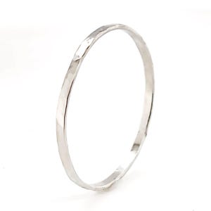 May include: A silver bangle bracelet with a hammered finish. The circular bracelet is made of metal and has a textured surface. The light reflects off the facets of the bracelet. The background is plain white.