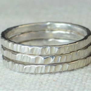 May include: Three silver rings stacked together. Each ring has a hammered texture, creating a unique, irregular surface. The rings are made of a shiny, metallic material, reflecting light.
