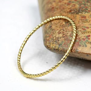 Puede incluir: Un brazalete de metal dorado retorcido. El brazalete tiene una textura similar a una cuerda y tiene forma ovalada. El brazalete descansa sobre una superficie marrón texturizada.