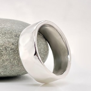 May include: A polished silver ring with a smooth, rounded design. The ring is resting on a gray stone, with a white background. The ring's surface reflects light, highlighting its shape and texture. This piece of jewelry is suitable for everyday wear.
