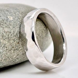 May include: A silver-colored ring with a hammered texture. The ring is set against a gray stone, showcasing its reflective surface. The ring's design is simple, yet elegant, suitable for everyday wear or special occasions.