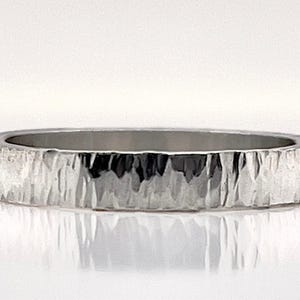 May include: A silver ring with a textured, bark-like surface. The ring has a flat profile and a polished interior. The design gives the ring a rustic, yet modern aesthetic. The ring is a band style.