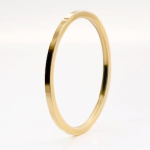 Könnte beinhalten: Ein dünner, goldfarbener Ring mit flachem Band. Der Ring ist vor einem weißen Hintergrund platziert, was sein schlichtes, elegantes Design hervorhebt. Die glatte Oberfläche des Rings reflektiert das Licht und erzeugt einen subtilen Glanz.