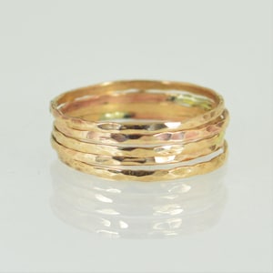 May include: A stack of six gold-colored hammered metal rings. The rings have a textured surface and reflect light, creating highlights and shadows. The rings are stacked on a reflective surface.