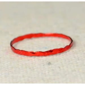 以下が含まれることがあります： テクスチャ加工された、ハンマー加工された外観の細い赤いリング。リングは鮮やかな赤色で、金属製のように見えます。リングは円形で、ニュートラルな質感の表面に置かれています。これはジュエリーです。