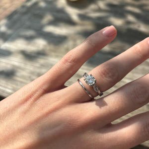Op de afbeelding: Een hand met twee zilveren ringen. De verlovingsring heeft een grote, ronde, heldere edelsteen in een gedraaide band. Daaronder zit een eenvoudige, dunne band. De ringen zitten aan een vinger met lichtroze nagellak.