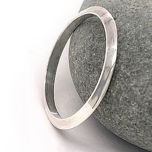 Pode incluir: Uma pulseira de bracelete prateada com um design minimalista. A pulseira redonda tem uma superfície lisa e polida. A pulseira tem aproximadamente 6,35 cm de diâmetro e repousa sobre uma pedra cinzenta.