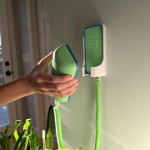 Puede incluir: Un dispositivo portátil verde y azul con un cable verde en espiral se sostiene cerca de un soporte de pared. El soporte es blanco con una inserción verde que tiene la palabra "TINER" impresa. El dispositivo es verde con ribetes azules.