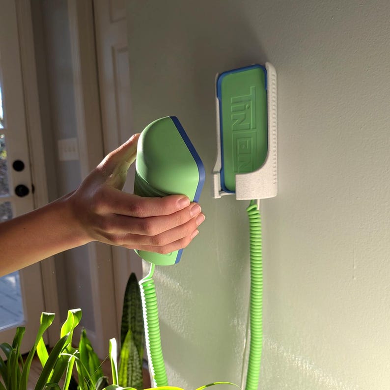 May include: A green and blue handheld device with a coiled green cord is being held near a wall-mounted holder. The holder is white with a green insert that has the word "TINER" printed on it. The device is green with blue trim.