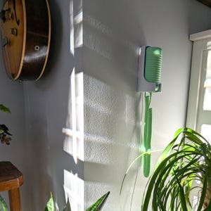 May include: A wall-mounted, retro-style telephone with a green handset and coiled cord. The phone is mounted on a white bracket against a gray wall. A vintage guitar hangs on the wall nearby, and a houseplant is in the foreground.