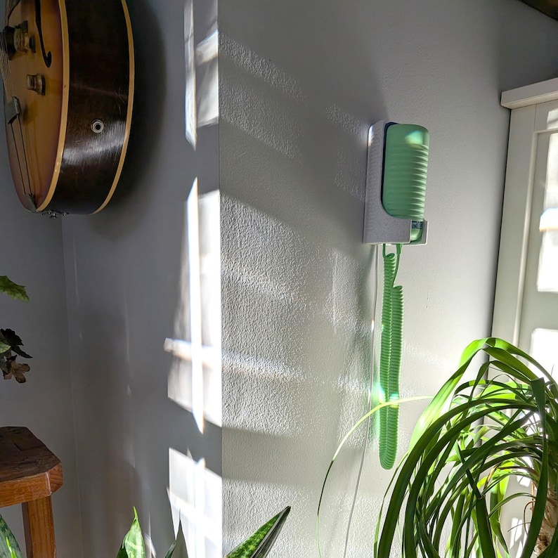 May include: A wall-mounted, retro-style telephone with a green handset and coiled cord. The phone is mounted on a white bracket against a gray wall. A vintage guitar hangs on the wall nearby, and a houseplant is in the foreground.