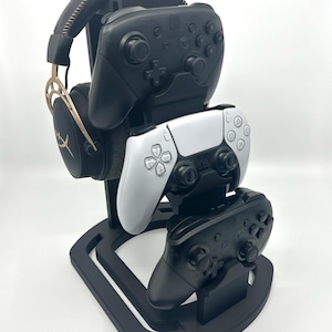 May include: Black gaming headset and three game controllers on a black stand. The controllers are black and white. The headset has gold accents.