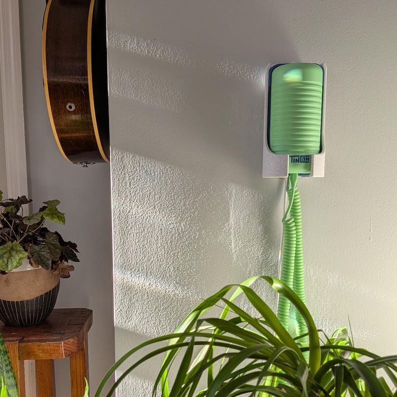 May include: A light green, cylindrical object mounted on a wall, with the text "TIN ON" visible. A green, coiled cord extends downward. A guitar and potted plant are also visible.
