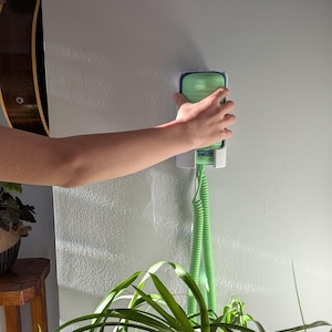 May include: A green and white wall-mounted plant mister with a coiled hose. The mister is being held by a person's hand. The word "LINCH" is visible on the mister. A guitar and a plant are in the background.