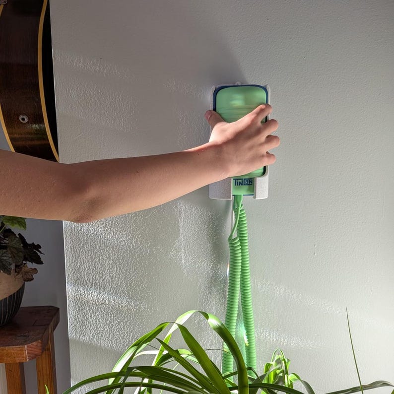 May include: A green and white wall-mounted plant mister with a coiled hose. The mister is being held by a person's hand. The word "LINCH" is visible on the mister. A guitar and a plant are in the background.