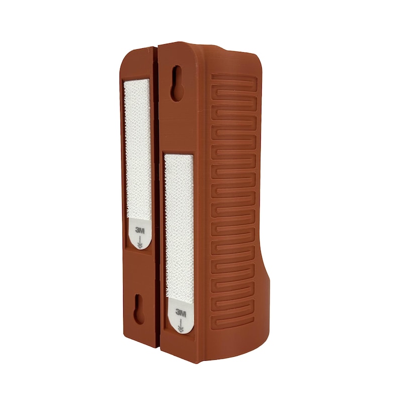 May include: A brown, rectangular wall mount with two sections. Each section has a white, rectangular adhesive strip with the "3M" logo. The right side has a series of vertical, indented lines. The mount has a curved bottom and a hole for hanging.