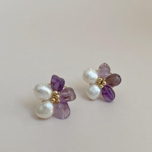 May include: A pair of floral earrings. Each earring is composed of white pearls and purple amethyst gemstones, arranged around a gold-coloured centre. The earrings are set against a light beige background.