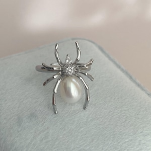 May include: A silver-colored ring featuring a spider design. The spider's body is a large, pear-shaped pearl, and its head is adorned with small, sparkling crystals. The spider's legs are outstretched, creating a unique and eye-catching piece of jewelry.