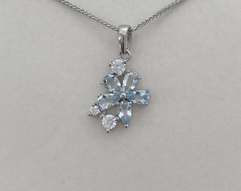 Puede incluir: Un collar con colgante plateado con un diseño floral. El colgante tiene piedras preciosas de color azul claro y transparente dispuestas en forma de flor. La cadena del collar es de color plateado delicado.