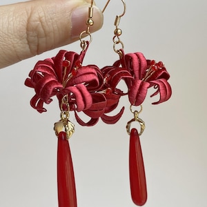 May include: A pair of red floral earrings with gold-colored hooks and accents. Each earring features a detailed, embroidered flower design and a long, teardrop-shaped red bead. The earrings are designed for pierced ears.
