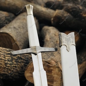 May include: A medieval-style sword with a white handle and scabbard. The sword features a silver crossguard and pommel, and the blade is silver. The scabbard is plain white with a decorative silver end. The sword is displayed against a backdrop of stacked logs.