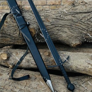 May include: A black sword with a silver crossguard and a black leather-wrapped handle. The sword is displayed next to its black sheath, which has a silver tip and a leather strap. The sword and sheath are resting on a pile of wooden logs.