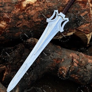 May include: A silver sword with a double-edged blade and a decorative hilt. The hilt is brown and wrapped, with a silver pommel. The sword is displayed on a textured, brown wooden surface.