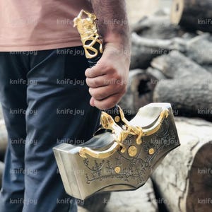 Puede incluir: Un martillo metálico detallado con detalles dorados y grabados intrincados. El mango es negro con un agarre dorado. El martillo se sostiene en la mano de una persona, con un fondo borroso de madera.