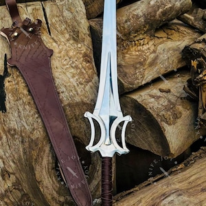 May include: A silver-colored sword with a decorative handle and a brown leather sheath. The sword has a wide blade and a detailed hilt. The sheath is brown and has a strap for carrying. The sword and sheath are displayed on a pile of wooden logs.