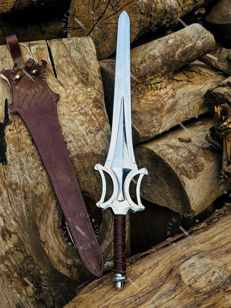 May include: A silver-colored sword with a decorative handle and a brown leather sheath. The sword has a wide blade and a detailed hilt. The sheath is brown and has a strap for carrying. The sword and sheath are displayed on a pile of wooden logs.