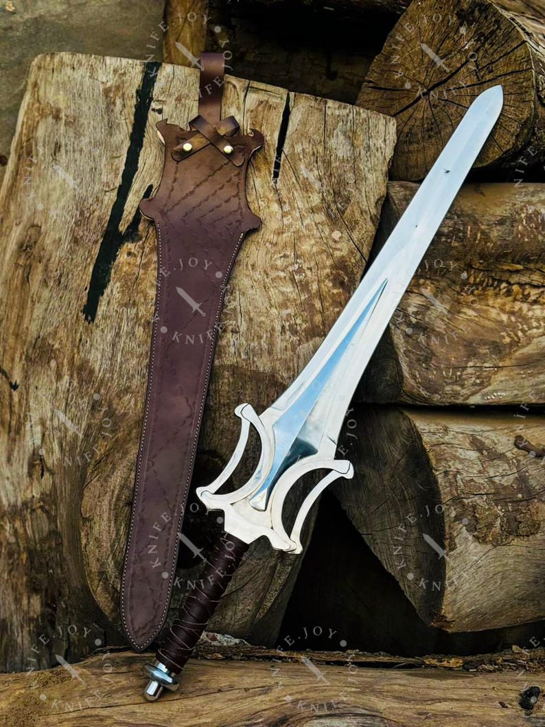 May include: A silver sword with a double-edged blade and a decorative hilt rests beside a brown leather sheath. The sheath is stitched and has a strap for carrying. The sword and sheath are displayed on a background of stacked wooden logs.