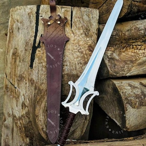 May include: A silver sword with a double-edged blade and a decorative hilt rests beside a brown leather sheath. The hilt is wrapped in dark brown leather. The sheath has a strap and is embossed with the word "KNIFE".