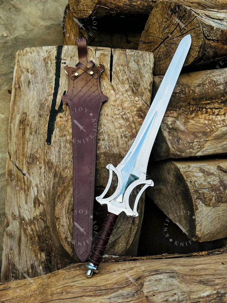 May include: A silver sword with a double-edged blade and a decorative hilt rests beside a brown leather sheath. The hilt is wrapped in dark brown leather. The sheath has a strap and is embossed with the word "KNIFE".