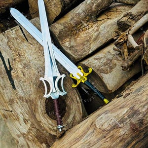 May include: Two decorative swords with silver blades and ornate handles rest on a pile of wooden logs. One sword has a silver and brown handle, while the other features a gold and black handle. The swords are designed with intricate details.
