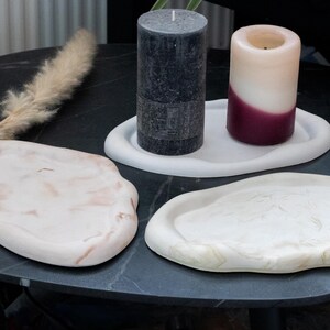 Peut inclure: Trois plateaux ovales de forme irrégulière avec des bords lisses et surélevés. Un plateau est blanc uni, tandis que les deux autres présentent des motifs marbrés dans des tons beige et crème. Une bougie pilier gris foncé et une bougie bicolore bordeaux et blanche reposent sur l'un des plateaux.
