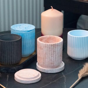 Peut inclure: Divers récipients cylindriques décoratifs et une bougie. Les récipients sont de différentes couleurs, dont le noir, le bleu et le rose, avec des détails côtelés verticaux. Un récipient a un couvercle. La bougie est de couleur pêche clair.