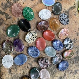 May include: A collection of various polished gemstones in oval shapes. The stones display a range of colors, including green, purple, blue, pink, and brown. Some stones have unique patterns and textures, such as stripes and speckles. The gemstones are displayed on a textured, brown surface.
