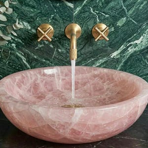 May include: A pink, oval-shaped stone sink with water flowing from a gold faucet. The sink is set against a green marble wall with gold fixtures. A gold vase with greenery and a glass soap dispenser are visible.