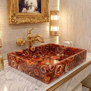 May include: A rectangular vessel sink crafted from polished red and brown agate stone slices, featuring a gold-toned metal faucet with two matching cross-handle knobs, set upon a white marble countertop with gray veining and a gold drain cover.