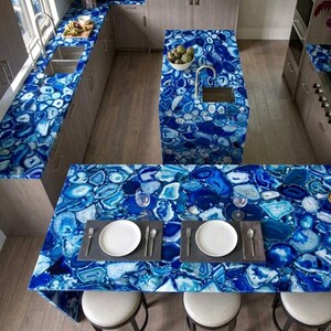 May include: A kitchen featuring countertops and a large island surfaced with polished blue agate stone slabs. The surfaces display a mosaic of deep blue, light blue, and white crystalline patterns. Two white plates sit on dark gray placemats.