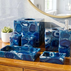 Può includere: Una collezione di accessori per il bagno in agata blu, tra cui un dosatore di sapone, un portacarte veline e varie scatole portaoggetti. I pezzi hanno una finitura lucida e sono disposti su una superficie di legno. È presente anche una piccola pianta succulenta.
