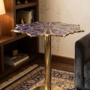 May include: A decorative side table featuring a purple, flower-shaped top. The tabletop is made of a marbled purple material with gold accents. The table has a gold-coloured, cylindrical stem and a round base. The table is placed on a patterned rug.