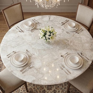 May include: A round dining table with a white marble top featuring a cracked stone pattern, set for four people with white ceramic plates, bowls, silver cutlery, and clear glassware. A glass vase with white lilies sits in the center.