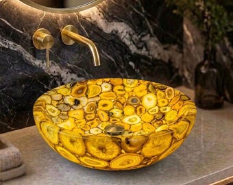 Może przedstawiać: Okrągła umywalka nablatowa wykonana z półprzezroczystych plastrów żółtego agatu, z polerowanym wykończeniem i centralnym metalowym odpływem. Umywalka prezentuje mozaikę naturalnych wzorów kamiennych w odcieniach złota, bursztynu i kremu na tle ciemnej marmurowej ściany.