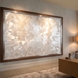 May include: Large rectangular wall art with a radiant, sunburst design. The artwork features a pattern of white, rectangular pieces arranged in a circular pattern. The artwork is framed with a dark wood and silver trim.