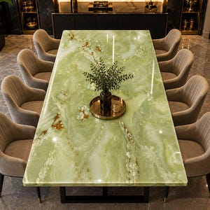 Peut inclure: Une table à manger rectangulaire avec un plateau en pierre d'onyx vert clair poli présentant des veines blanches et dorées, reposant sur une base en métal noir. Dix chaises taupe rembourrées à dossier cannelé entourent la table.