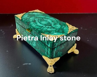 May include: A rectangular, green malachite box with gold-coloured accents and feet. The box features a swirling pattern and the text "Pietra Inlay stone" in white. The corners and feet are adorned with ornate gold detailing.