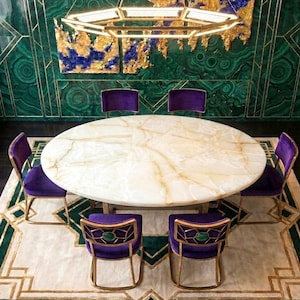 Op de afbeelding: Een grote ronde eettafel van witte steen met gouden aders, omringd door zes paarse fluwelen stoelen met gouden metalen frames. De rugleuningen hebben een geometrisch goudpatroon met een ronde groene steen in het midden.