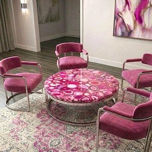 May include: A round coffee table with a polished pink agate stone top and a silver metal frame, surrounded by four matching pink velvet armchairs with silver metal frames, all positioned on a patterned rug with pink and grey tones.