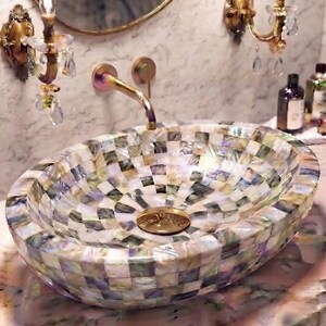May include: An oval vessel sink featuring a mosaic pattern of iridescent, multi-colored mother-of-pearl tiles. The sink includes a polished gold-tone drain cover and is paired with a wall-mounted, curved gold-tone faucet fixture.